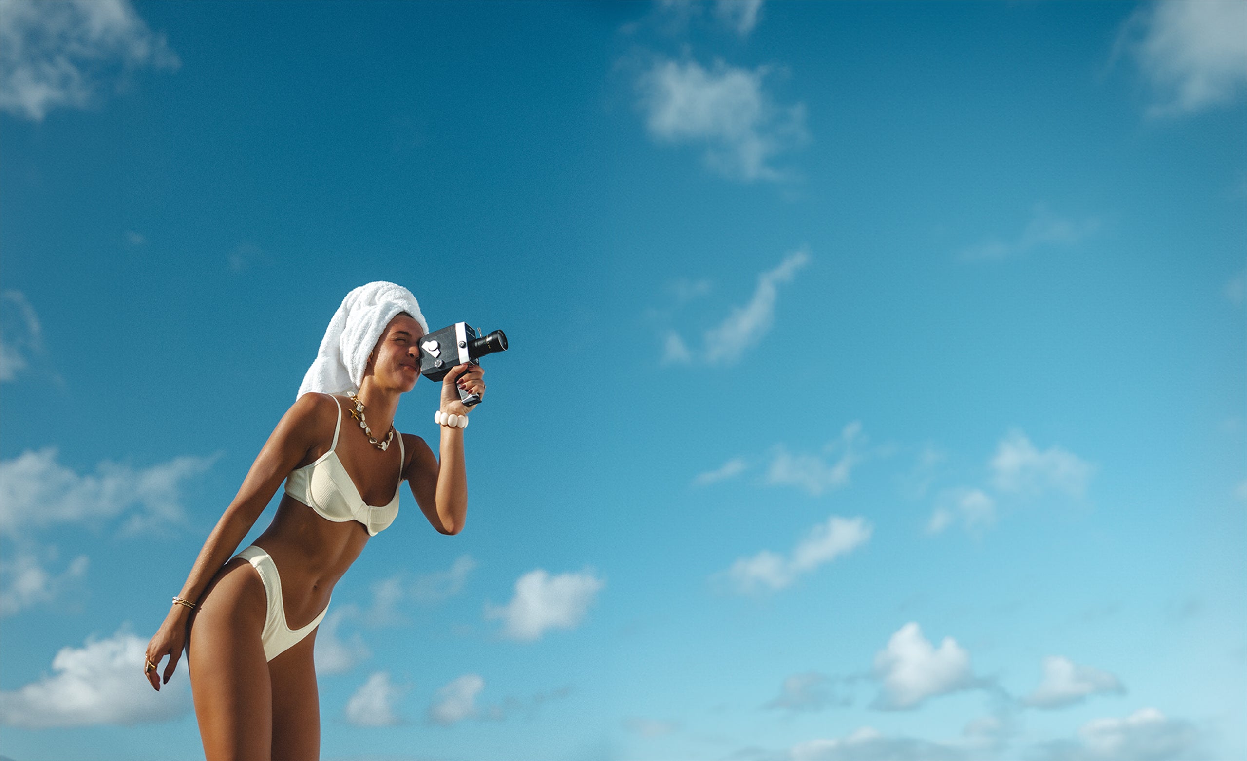 A person in a white bikini and a towel turban uses a vintage camera against a bright blue sky with scattered clouds. They are smiling and appear relaxed, capturing a moment in the open air.