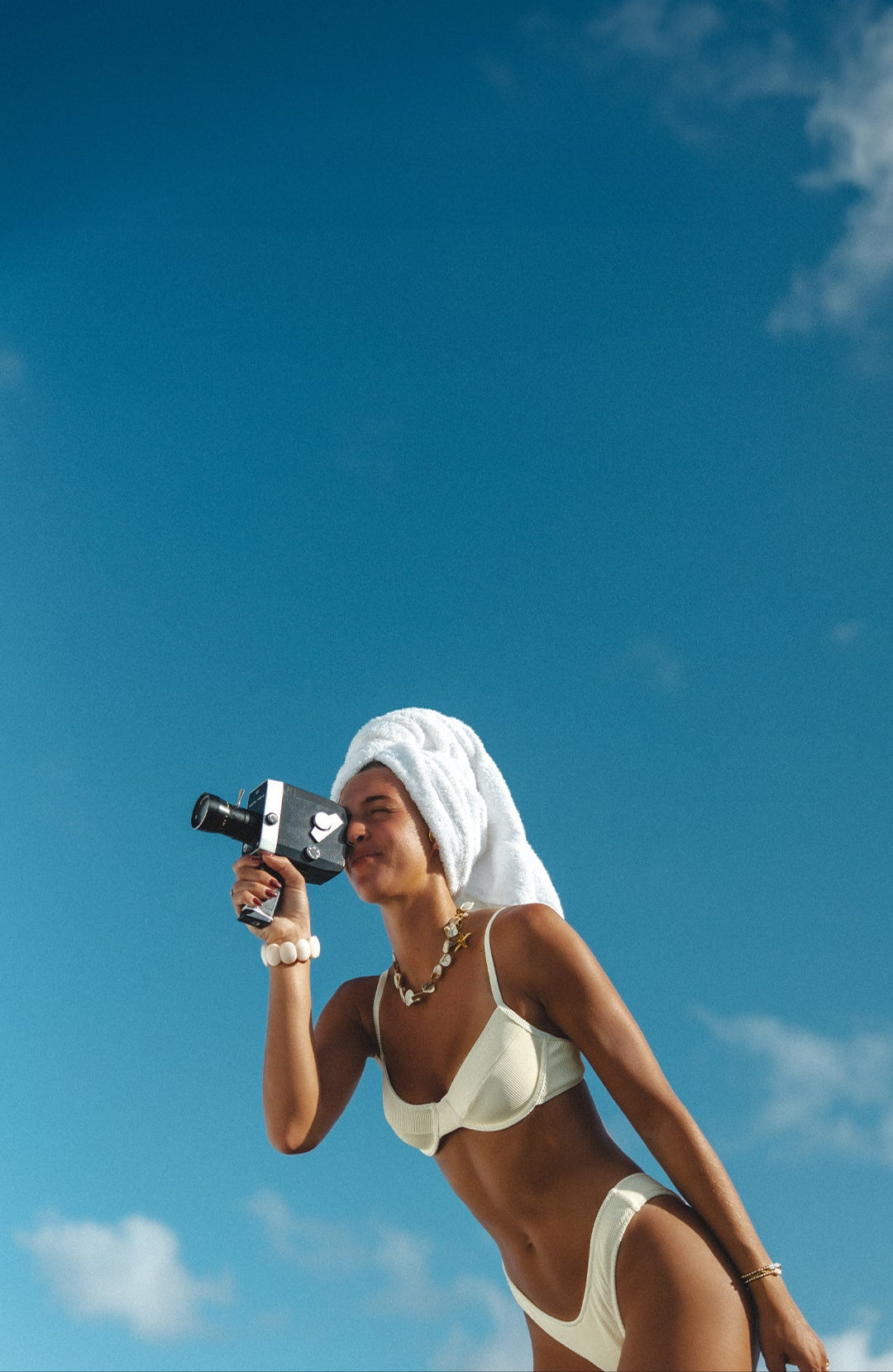 A woman in a white bikini with a towel wrapped around her head is smiling and holding a vintage video camera. The sky behind her is clear and blue. She appears relaxed and content in the sunny setting.